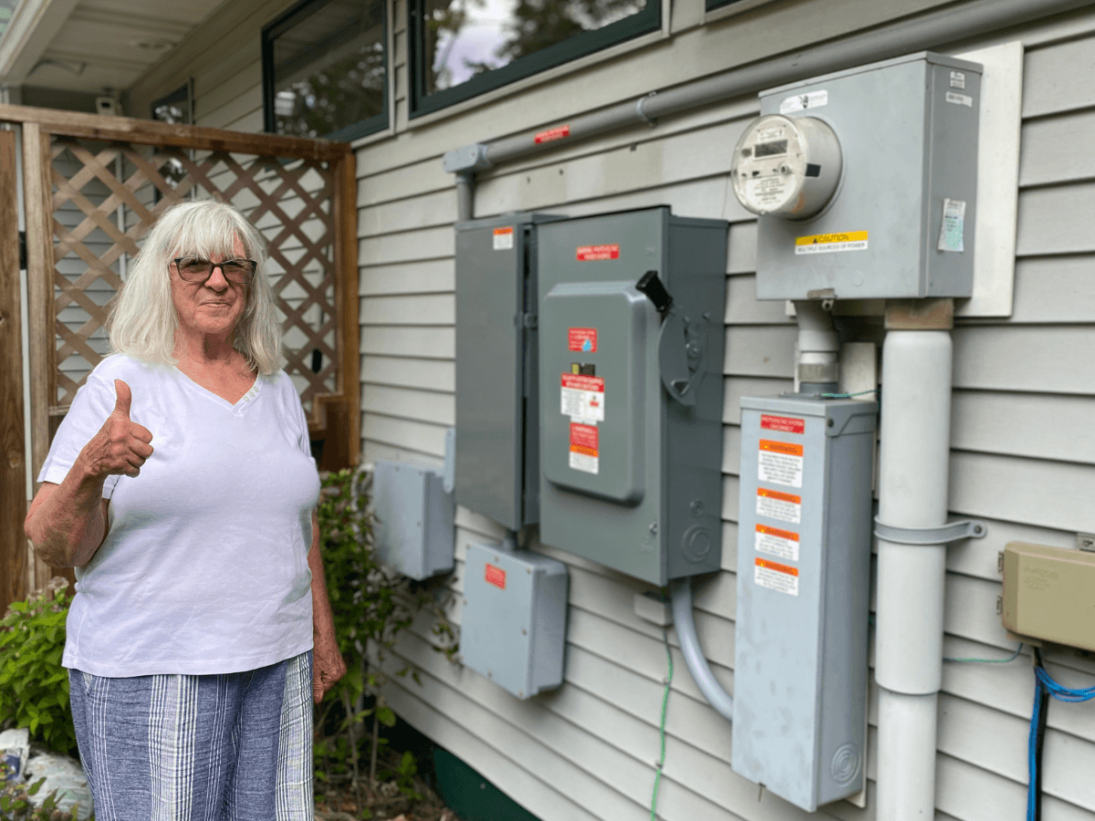 A smiling woman gives a thumbs up next to electrical equipment mounted on a house wall.